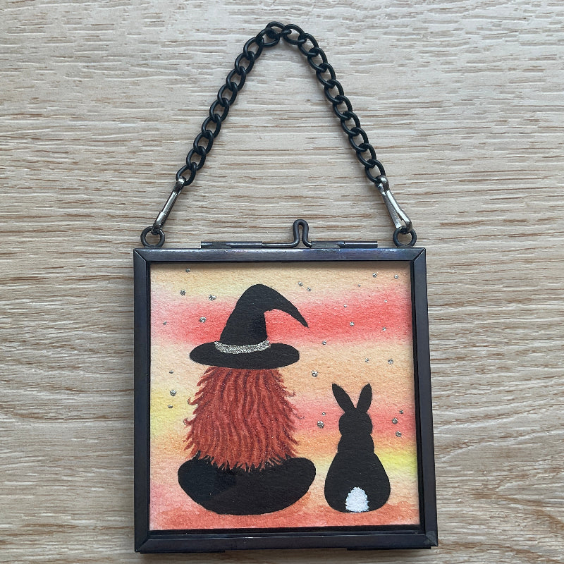 Original mini watercolor painting titled Me and My Shadow featuring a witch and a black rabbit silhouette against a vibrant orange sunset background with silver stardust.