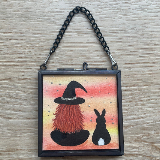 Original mini watercolor painting titled Me and My Shadow featuring a witch and a black rabbit silhouette against a vibrant orange sunset background with silver stardust.