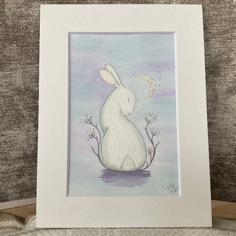 Original watercolor painting titled Moonbeam Kisses featuring a white rabbit looking up at a crescent moon with silver highlights.