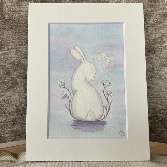 Original watercolor painting titled Moonbeam Kisses featuring a white rabbit looking up at a crescent moon with silver highlights.