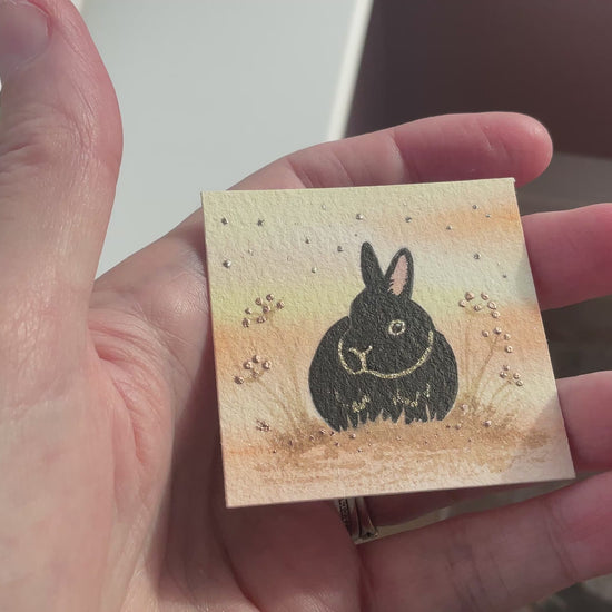 Video of Original mini watercolor painting titled Autumn Whispers featuring a black rabbit silhouette with gold outlines and silver metallic stardust in a hanging metal frame
