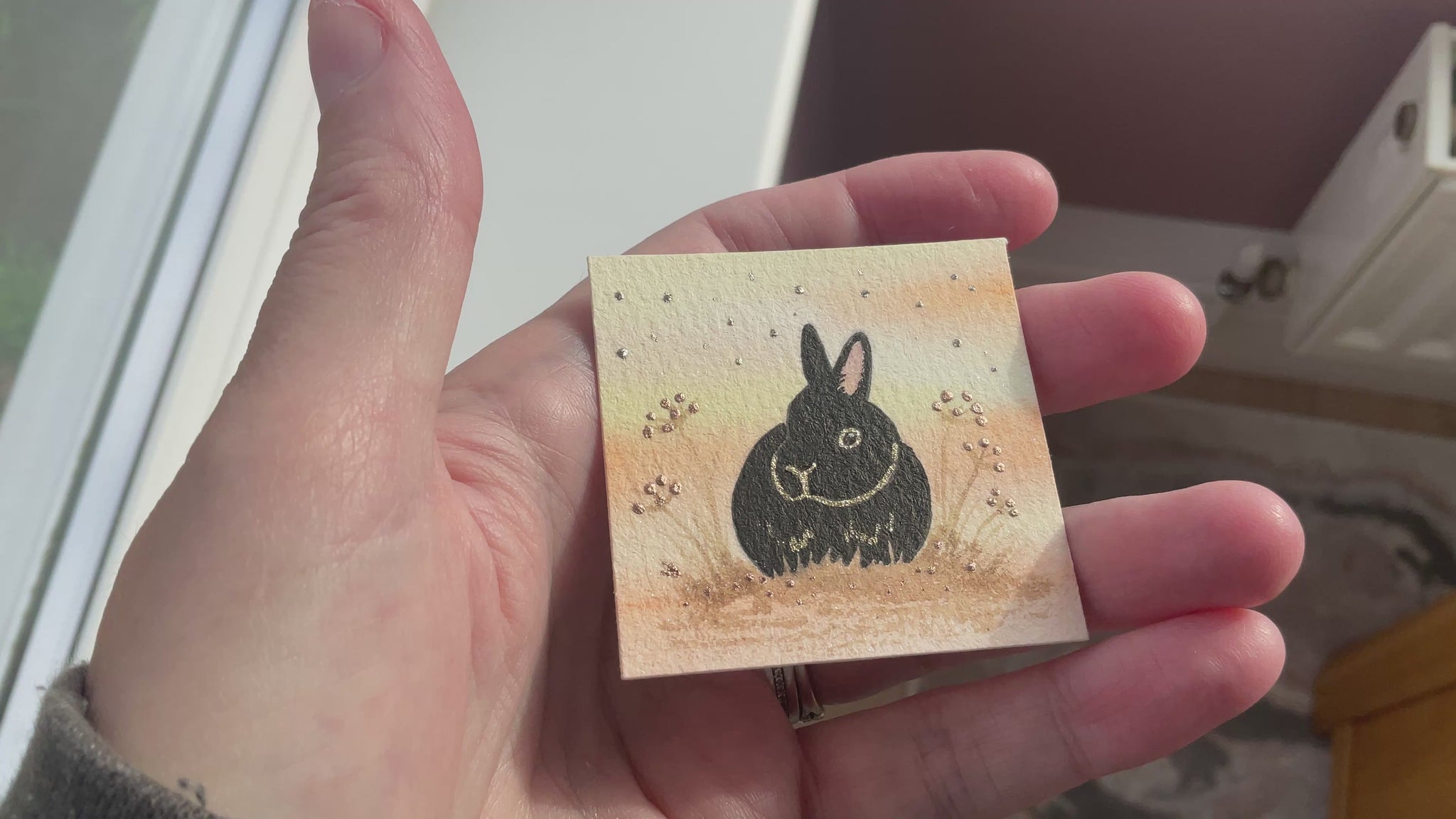 Video of Original mini watercolor painting titled Autumn Whispers featuring a black rabbit silhouette with gold outlines and silver metallic stardust in a hanging metal frame