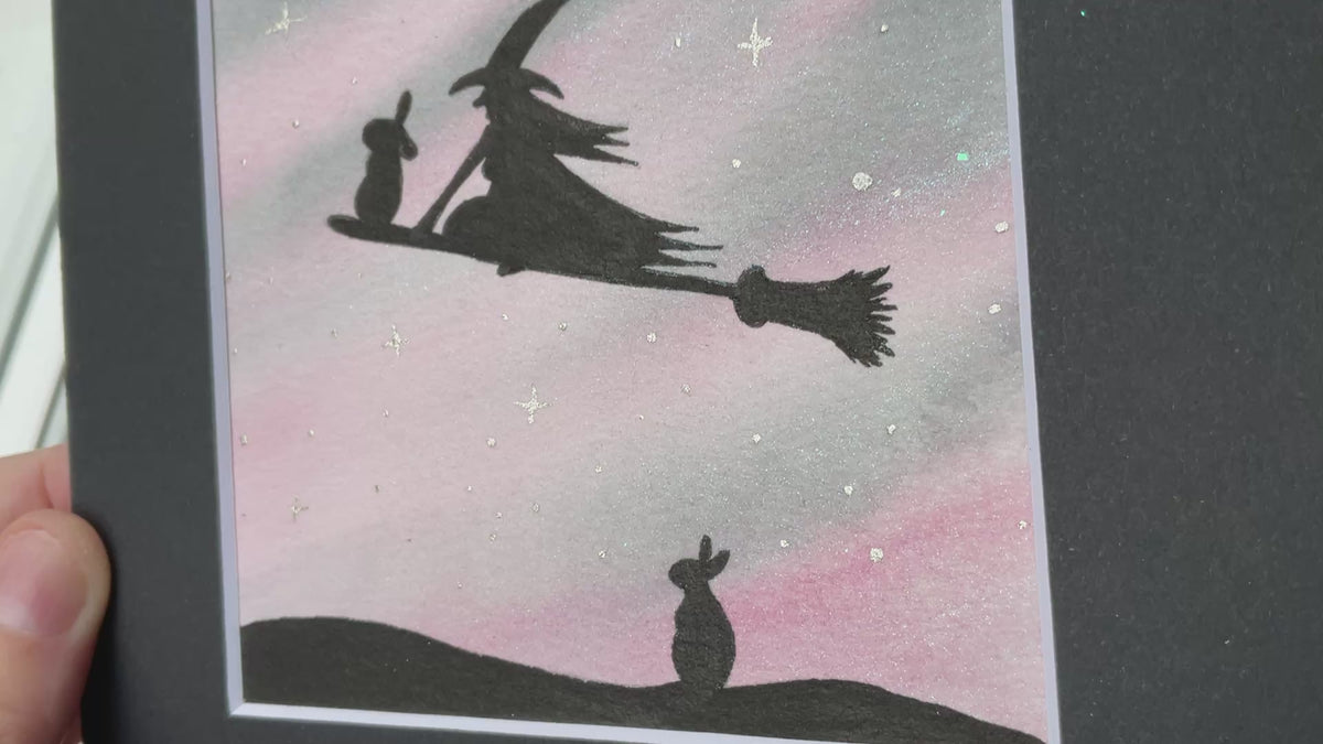 Video Original mini watercolor painting titled Waiting My Turn featuring a witch and a rabbit flying on a broomstick while a second rabbit waits on the ground under silver stars.