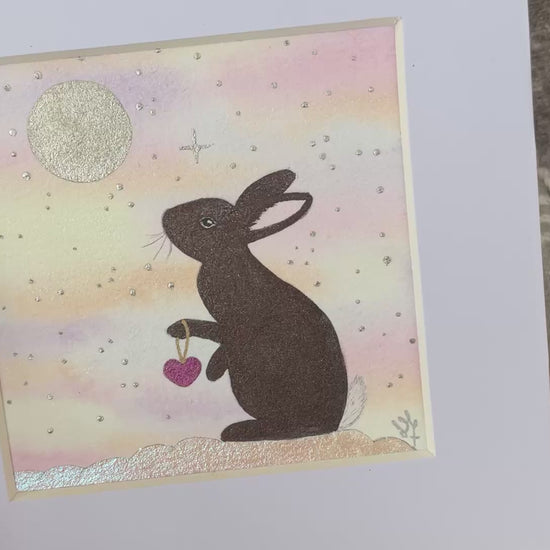 Video of Comforting original brown bunny painting holding a pink heart under a full moon and rainbow skies mounted in a white frame
