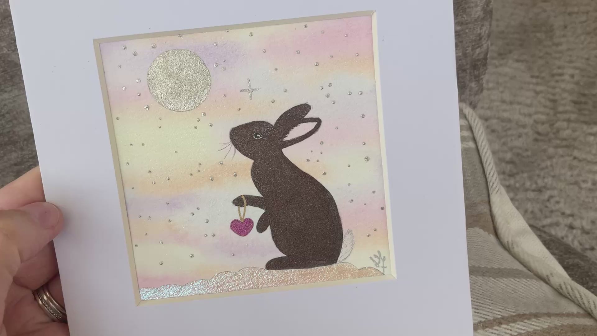 Video of Comforting original brown bunny painting holding a pink heart under a full moon and rainbow skies mounted in a white frame