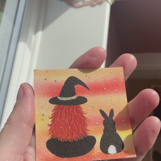 Video of Original mini watercolor painting titled Me and My Shadow featuring a witch and a black rabbit silhouette against a vibrant orange sunset background with silver stardust.