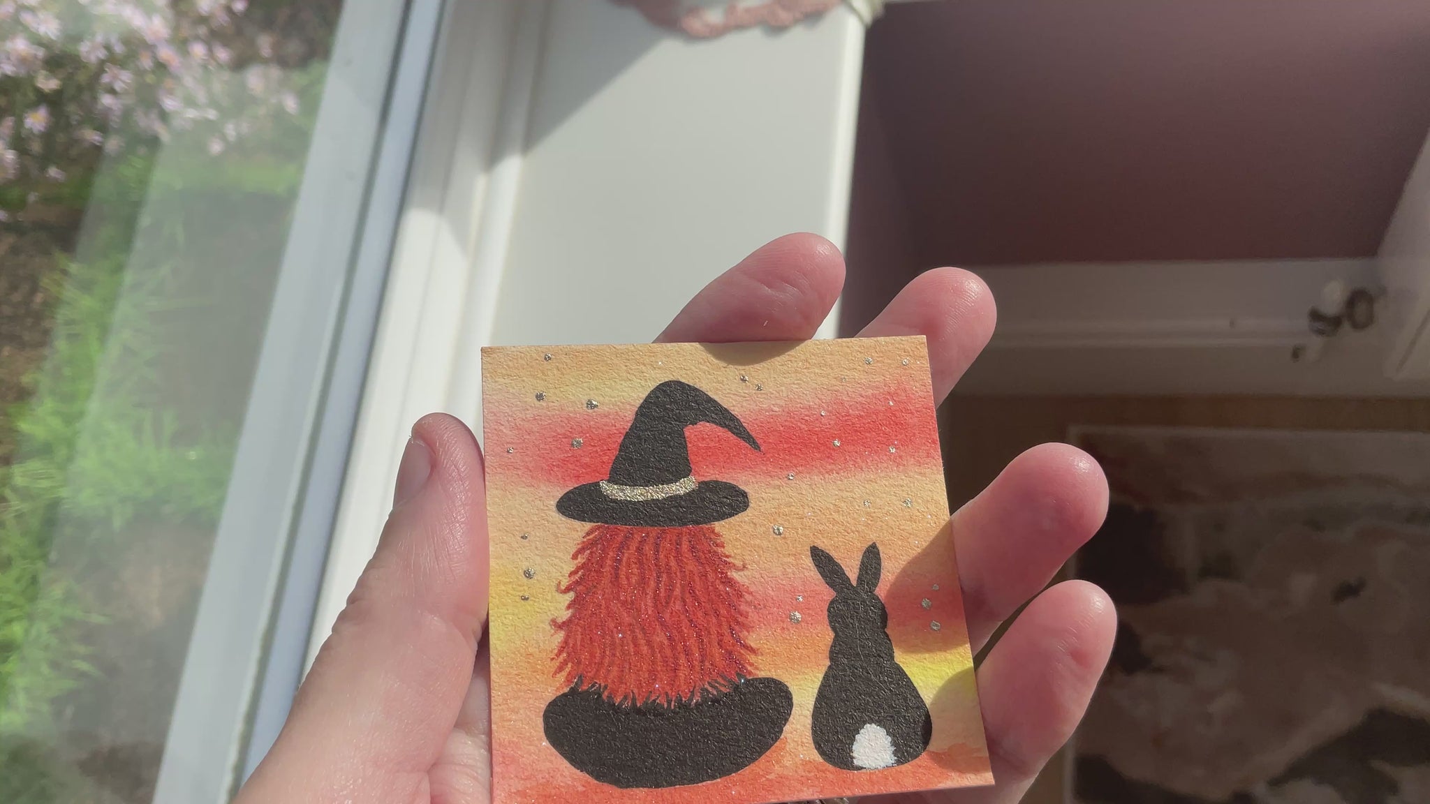 Video of Original mini watercolor painting titled Me and My Shadow featuring a witch and a black rabbit silhouette against a vibrant orange sunset background with silver stardust.