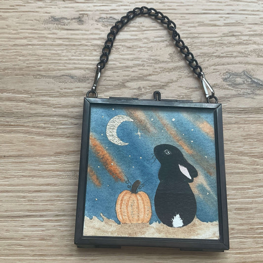 Stardust and Pumpkins autumn watercolor painting showing a black bunny with a white tail next to a glowing pumpkin under a crescent moon