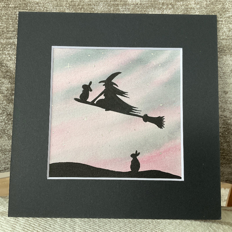 Original mini watercolor painting titled Waiting My Turn featuring a witch and a rabbit flying on a broomstick while a second rabbit waits on the ground under silver stars.